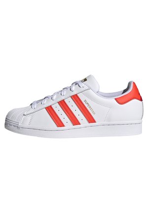 Białe niskie sneakersy z czerwonymi paskami i pętelką na pięcie, ochronnym noskiem, sznurowanym fasonem oraz napisem „Superstar” po boku.