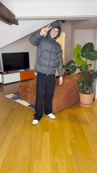 Giacca imbottita grigia con cappuccio, pantaloni neri e scarpe bianche. Ambiente interno con divano marrone, grande pianta verde e pavimento in legno.