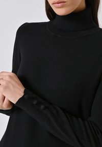 Frau in einem schwarzen Rollkragenpullover mit drei stoffbedeckten Knöpfen am Bündchen, die Hände nahe dem Handgelenk gefaltet.