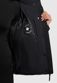 Černá bunda se hladkou vnitřní podšívkou, s logem, patentkami a kapsou na zip. Design zahrnuje přiléhavý střih a delší délku.
