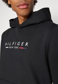 Asmuo dėvi juodą gobtuvą su užrašu „HILFIGER NEW YORK“ ir raudonomis, baltomis bei mėlynomis detalėmis krūtinės srityje.