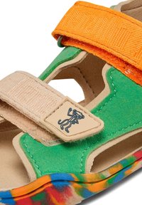 Sándala multicolor con parte superior de ante verde, tiras naranjas y beige, y una suela con un patrón colorido que presenta un emblema de personaje en la tira.