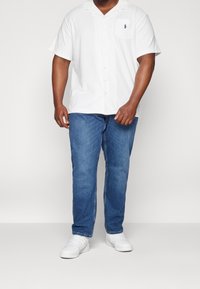 Camicia bianca a maniche corte con bottoni e taschino sul petto, abbinata a jeans blu e sneakers bianche. La camicia presenta un logo discreto.