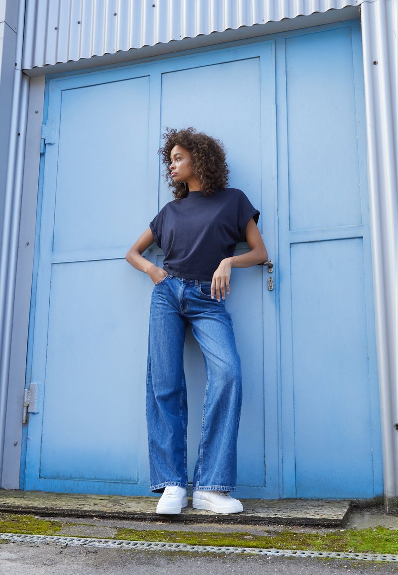 Marineblaues Kurzarm-Oberteil, hochtaillierte weit geschnittene blaue Jeans und weiße Sneaker. Hintergrund: hellblaue Holztür mit horizontalen Linien.