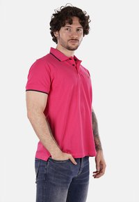 Polo shirt rosa con colletto classico, accentato in blu navy, e maniche corte. Ricamo visibile sul petto. Indossato con jeans.