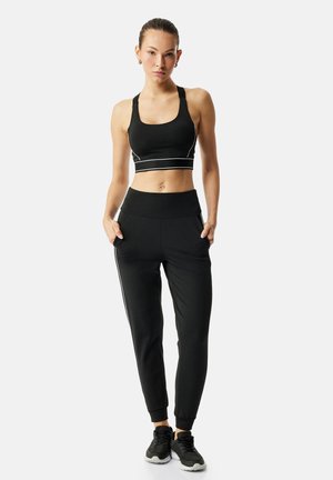 Femme debout portant un soutien-gorge de sport noir, un pantalon de jogging noir avec des bandes latérales et des chaussures de sport noires, mains dans les poches, fond neutre.