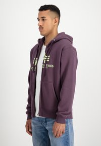 Fialová mikina na zips s predným vreckom a šnúrkou na kapucni. S kontrastným žltým logom na hrudi a príjemne pruhovanými manžetami a spodným lemom.