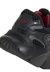 Black athletic shoe featuring a mesh upper with red accents, rubber sole, textured leather overlays, and a prominent logo on the side.