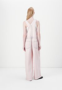 Rosa denim jumpsuits med vide ben, justerbare stropper og store baklommer. Minimal sømdetalj skaper kontrast til teksturen i stoffet.