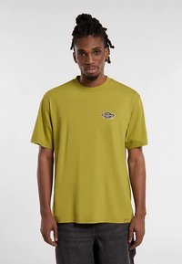 Oliwkowa bawełniana koszulka z okrągłym dekoltem, krótkimi rękawami oraz małym czarno-białym logo na lewej piersi.