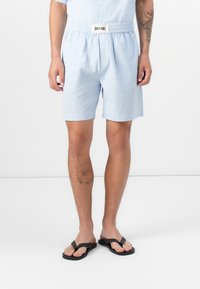 Hellblaue Baumwollshorts mit elastischem Bund und einem Logofähnchen. Der Stoff hat ein strukturiertes Muster; kombiniert mit schwarzen Flip-Flops.