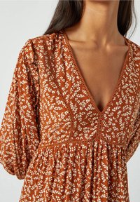 Donna che indossa un vestito lungo arancione ruggine a maniche lunghe con un motivo floreale bianco e scollatura a V profonda con dettagli decorativi.