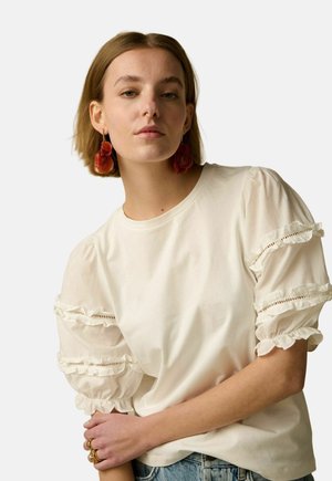 Jeune femme aux cheveux courts portant un chemisier crème à manches bouffantes à volants et de grandes boucles d'oreilles rouges en forme de fleur.