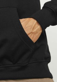 Sudadera negra con un gran bolsillo frontal, puños de canalé y una textura suave. Mano colocada en el bolsillo, mostrando detalles de la piel.