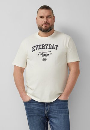 Man met kort haar en baard, draagt een off-white "EVERYDAY Rethink" t-shirt en blauwe jeans, handen in de zakken, staand tegen een grijze muur.
