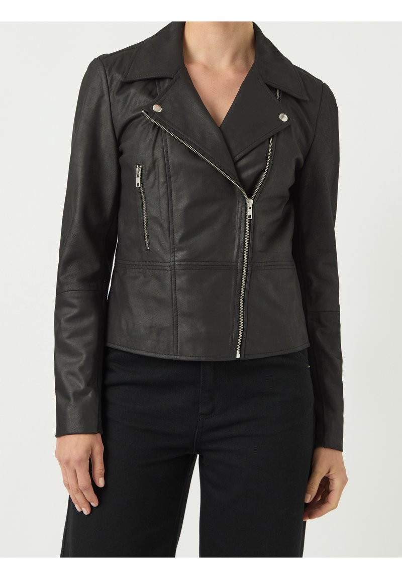 Femme portant une veste en cuir noire de motard avec une fermeture éclair argentée asymétrique et une poche poitrine zippée, associée à un pantalon noir.