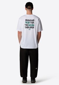 T-shirt bianco con testo nero e teal sul retro che dice "FORNIRE ATTREZZATURA PER L'ESPLORATORE MODERNO." Pantaloni neri e scarpe.