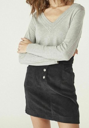 Kobieta ubrana w jasnoszary sweter z dekoltem w serek z misternym wzorem dzianiny oraz czarną mini spódnicę sztruksową z dwoma guzikami i boczną kieszenią.