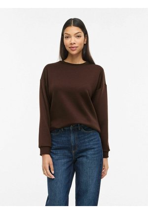 Noor naine, kellel on pikad tumedad juuksed, pruun pika varrukaga kampsun ja sinised teksad, seisab ühevärvilise valge tausta ees.