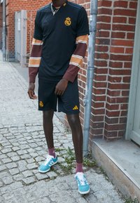 Černý dres s žlutým emblémem a pruhovanými rukávy v hnědé a béžové barvě, spárovaný s černými šortkami s logem. Tyrkysové tenisky s bílými akcenty.