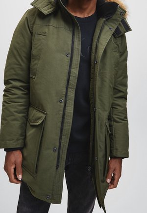 Homme portant une veste parka vert olive avec plusieurs poches, fermeture éclair noire, boutons-pression et capuche bordée de fourrure, partiellement ouverte sur une chemise noire.