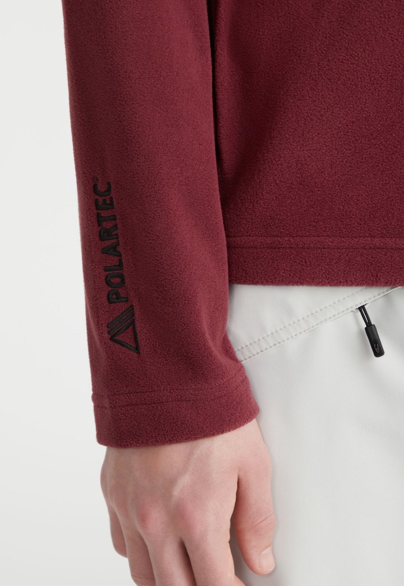 Pull-over en polaire bordeaux avec la marque Polartec en noir sur le poignet. Présente une surface texturée et un ourlet ajustable avec un cordon de serrage.