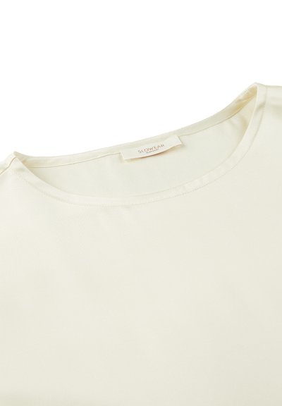 Cremefarbenes, kurzärmliges Oberteil aus glattem Stoff mit Rundhalsausschnitt. Verfügt über ein Etikett am Hals mit der Aufschrift "SLOWEAR".