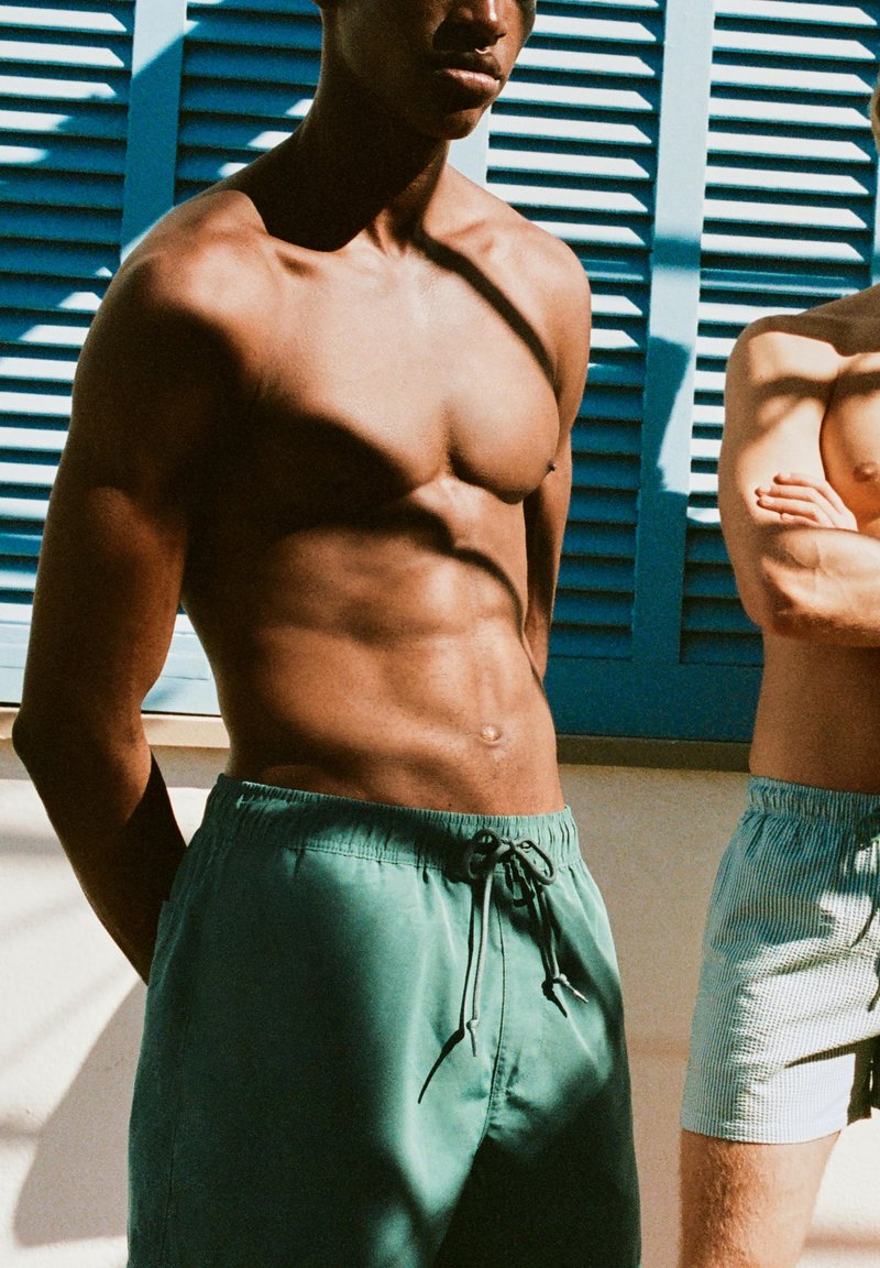 Deux hommes torse nu en shorts de bain se tiennent devant des fenêtres à volets bleus, avec la lumière du soleil projetant des ombres sur eux.