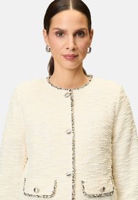 Texturierte cremefarbene Jacke mit schwarzem Besatz, rundem Halsausschnitt und silbernen Knöpfen. Verfügt über zwei frontale Taschen mit Knopfakzenten.