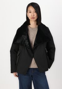 Black puffer jacket with a glossy lapel, featuring a fitted design and two side pockets. Worn over a beige textured sweater and blue jeans.