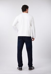 Λευκό πουκάμισο με μακριά μανίκια, σκούρα μπλε τζιν παντελόνια, μαύρα και λευκά αθλητικά παπούτσια. Ομαλό ύφασμα, κανονική εφαρμογή, τυπικό κόψιμο. Θέα από πίσω.