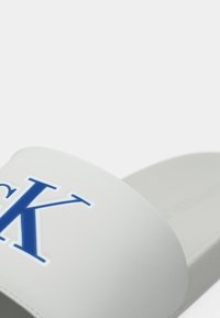 Közelkép egy fehér gumiszandálról, amelynek pántján nagy kék "CK" felirat található, fehér háttér előtt.