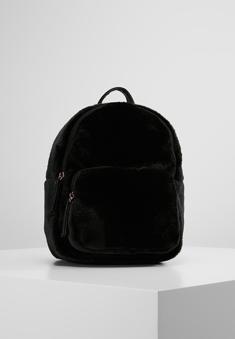 Sac à dos en peluche noire avec poche zippée à l'avant et poignée supérieure, posé sur une surface blanche devant un fond gris uni.