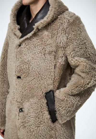 Beżowy płaszcz z kożuchem z dużym kapturem, skórzanymi akcentami na kołnierzu i kieszeniach, wykonany z teksturowanej tkaniny z zapięciem na guziki.