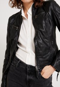 Veste de moto en cuir noir avec poches zippées, design ajusté et finition texturée, portée sur une chemise blanche à boutons et un jean foncé.