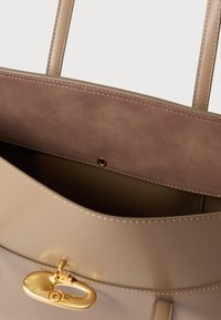 Beige bőr kézitáska sima textúrával, arany csattal és sötétbarna belső béléssel. Két fogantyúval és varrott részletezéssel kialakítva.