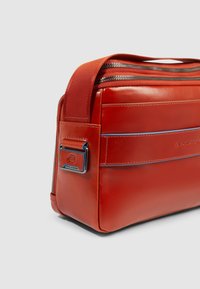 Bolso tipo mensajero de cuero rojo con cierre de cremallera, textura lisa, líneas de acento azules y una placa de logotipo metálico en el lateral. Forma cuadrada y correa resistente.