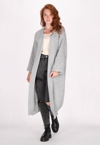 Lang grijs gebreide cardigan met open voorkant, gestructureerd patroon, brede mouwen, gedragen over een beige top en versleten zwarte jeans, gecombineerd met zwarte laarzen.