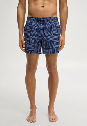 Man draagt blauw-zwarte zwemshorts met een geometrisch patroon, staat op blote voeten tegen een effen lichte achtergrond.