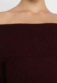 Pull en tricot bordeaux à épaules dénudées, avec texture côtelée, un col large et des manches longues avec un corps ajusté.