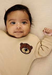 Smėlio spalvos ilgarankoviai bodžiukas su siuvinėta rudai kreminės spalvos meškos veido aplikacija ir spaudėmis ant peties. Fonas – šviesi medžiaga.