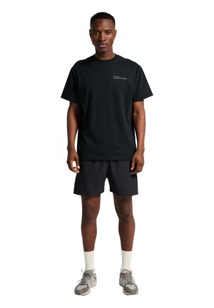 Mand står vendt fremad iført sort t-shirt med lille hvid tekst, sorte shorts, hvide sokker og grå sneakers på hvid baggrund.