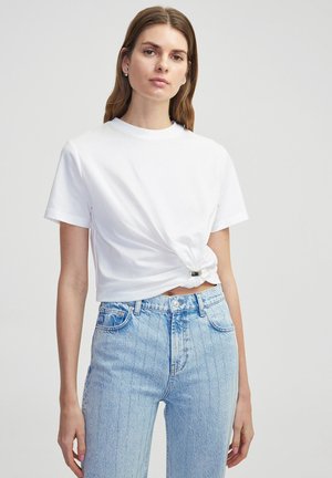 Camiseta blanca recortada con un detalle de nudo retorcido en la cintura, combinada con jeans de mezclilla azul claro con rayas verticales y una bragueta de botones.