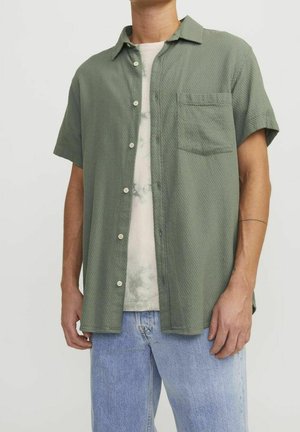 Mężczyzna noszący jasną zieloną koszulę z krótkim rękawem zapinaną na guziki na jasnym tie-dye T-shircie i jasnoniebieskich dżinsach, stojący na jednolitym tle.