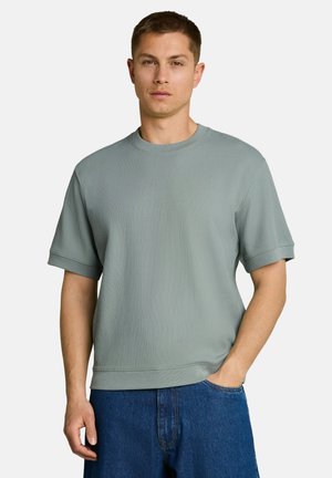 Jonge man met kort bruin haar, gekleed in een lichtgroen geribd T-shirt met korte mouwen en een blauwe spijkerbroek, staand met één hand in de zak.