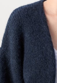 Námořnický pletený cardigan s texturovaným, načechraným povrchem a otevřeným předním dílem. Měkká tkanina volně splývá kolem ramen.
