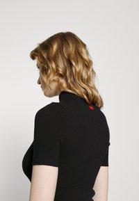 Černý žebrovaný top s krátkým rukávem a vysokým límcem, který má vzadu malou červenou značku. Měkčený materiál s přiléhavým střihem.