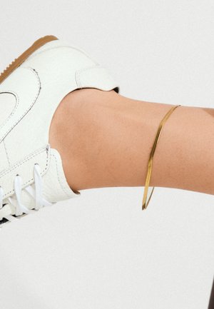 Sneaker blanche avec des lacets noirs, accompagnée d'un bracelet jonc doré mince sur une cheville nue. Texture lisse et design minimaliste.