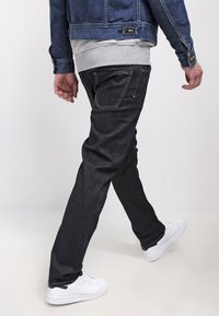Jeans de mezclilla oscura con corte recto, que presentan costuras visibles, bolsillos frontales y un aspecto desgastado. Combinados con una sudadera gris y zapatillas blancas.