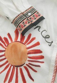 Wit shirt met een groot oranje zonontwerp met rode stralen, aangevuld met een gestreepte manchetten in zwart en beige met bloemaccenten.